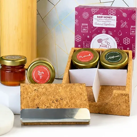 Four small raw honey jars, two in a tray and two beside it, in front of the printed pink Raw Honey box