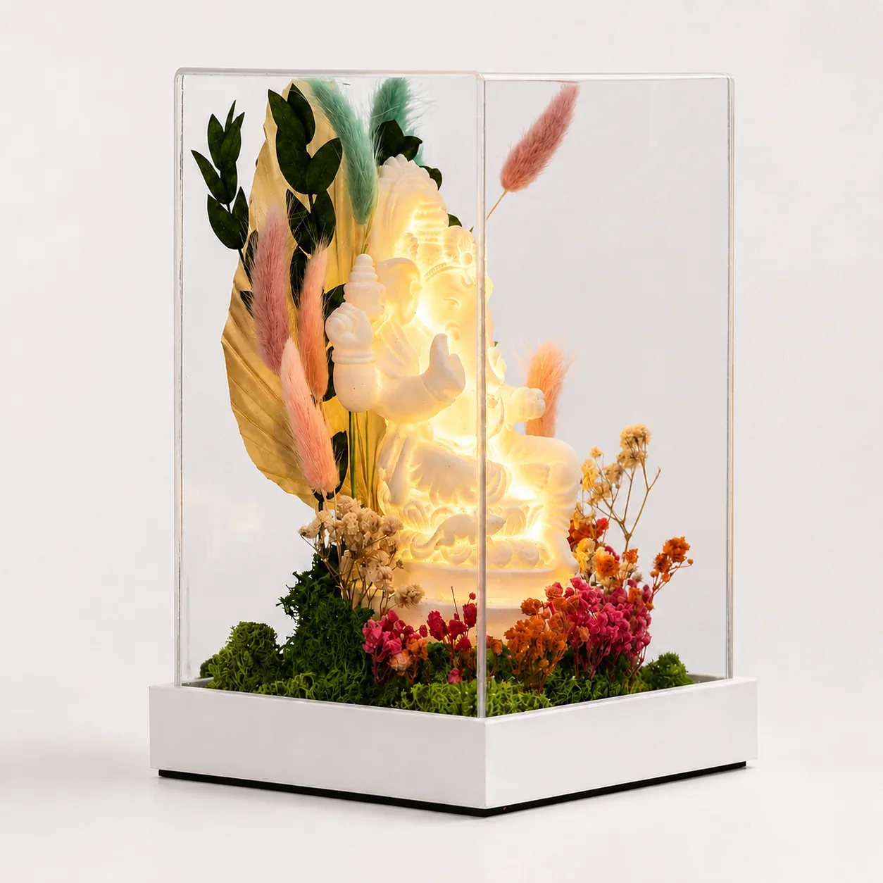 Forest Terrarium Ganesha, lit, three-quarter view: a glowing white Ganesha with preserved flowers in a tall clear acrylic cover on a white base