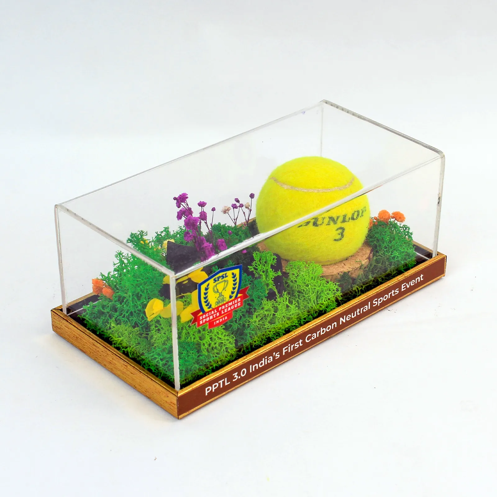A yellow tennis ball set in preserved moss and dried flowers under a clear acrylic cover, on a wooden base printed with the tournament's line
