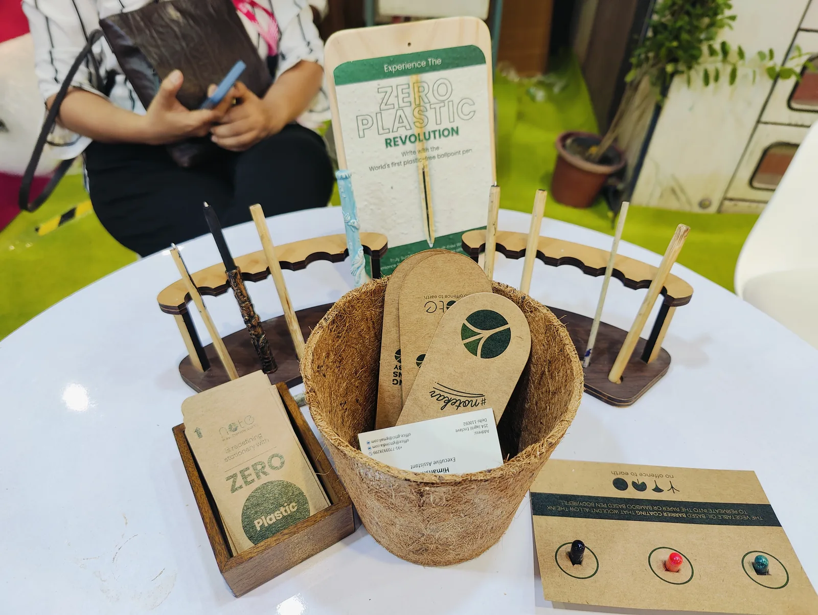 A white table with a coir basket of cards, pens in a wooden stand and a card reading Zero Plastic Revolution