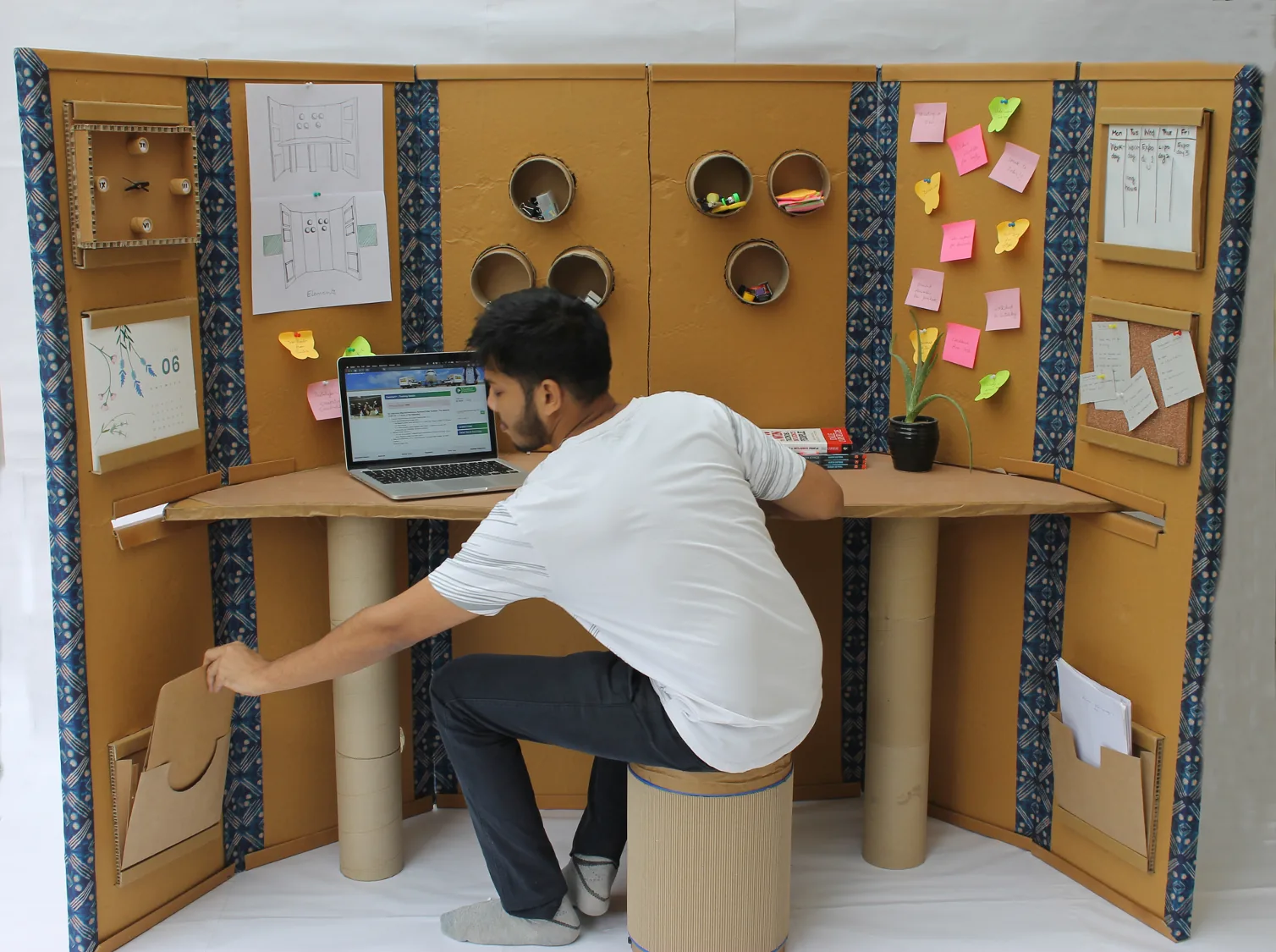 A person working at a standing cardboard desk with a laptop, a stool and pinboards, on a white floor