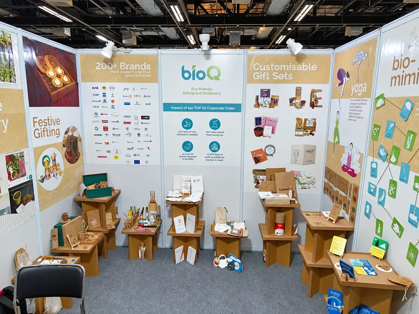 A stall with white panels and cardboard stools holding notebooks and gifts, with a wall of brand logos