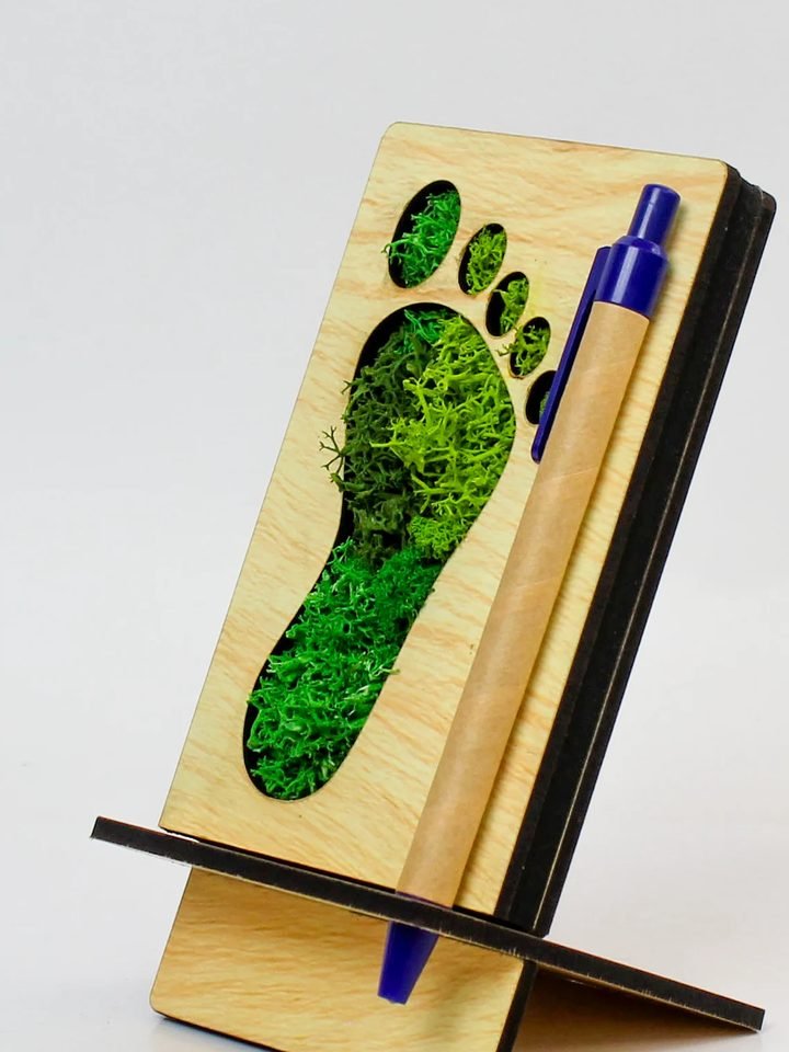 A wooden desk stand with a footprint cut out of it, moss showing through, and a pen at its side